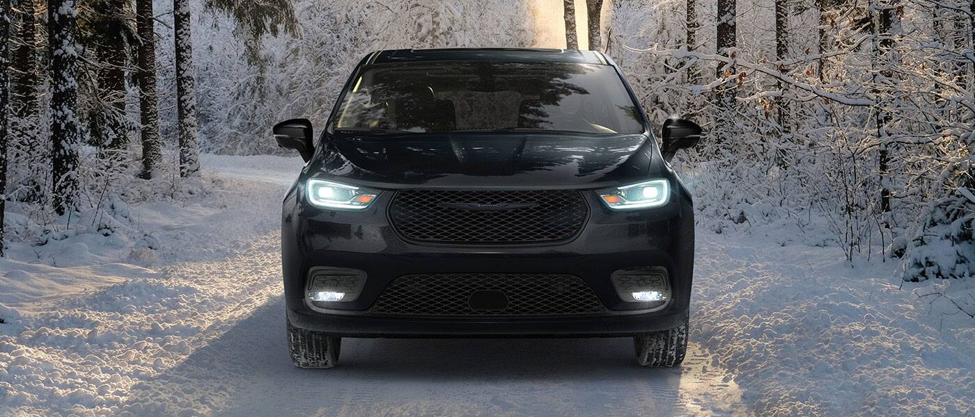 A head-on angle of a black 2026 Chrysler Pacifica Limited with its headlamps on, traveling down a snow-covered road in the woods.