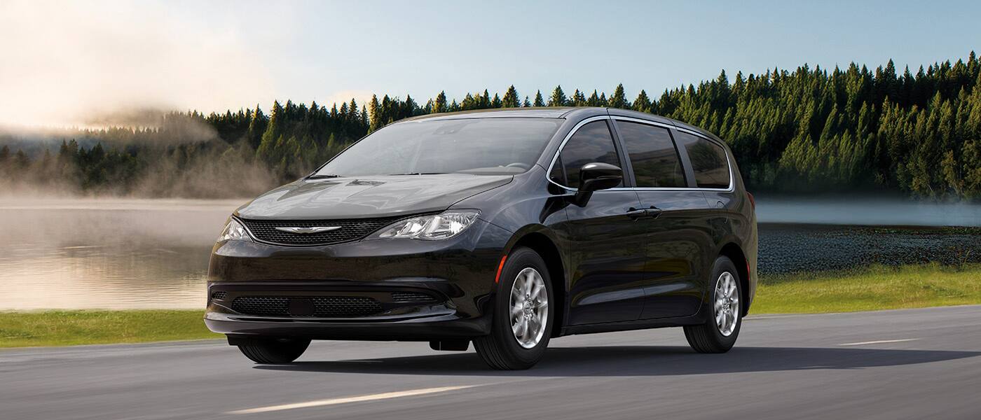 A driver-side front angle of a black 2026 Chrysler Voyager LX traveling on a lakeside highway.
