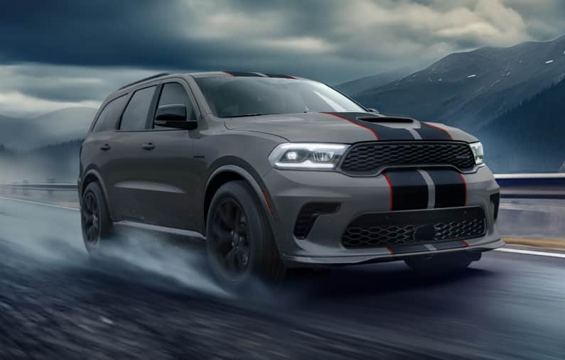 A gray 2026 Dodge Durango SRT Hellcat Jailbreak with black dual center stripes outlined in red, traveling down a highway in the rain with storm clouds overhead and mountains in the distance.