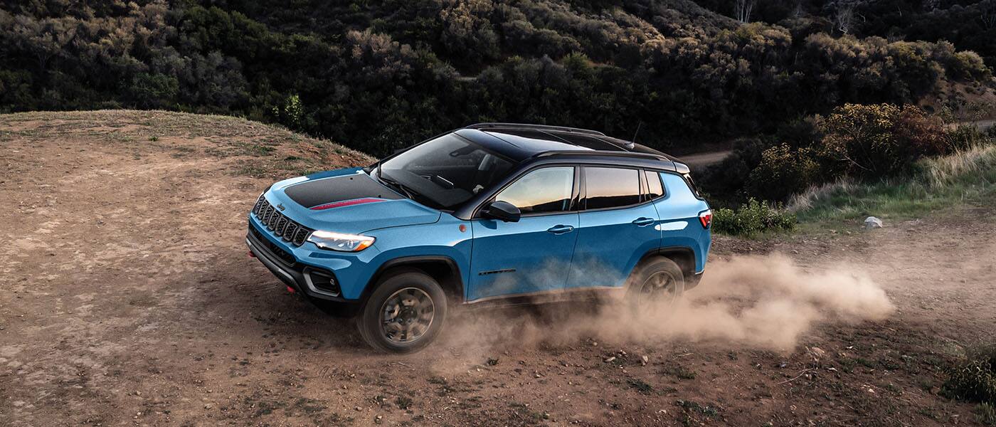 A blue 2026 Jeep Compass Trailhawk with a black hood insert and black roof, ascending a hill on a trail off-road, with the tires kicking up dust.