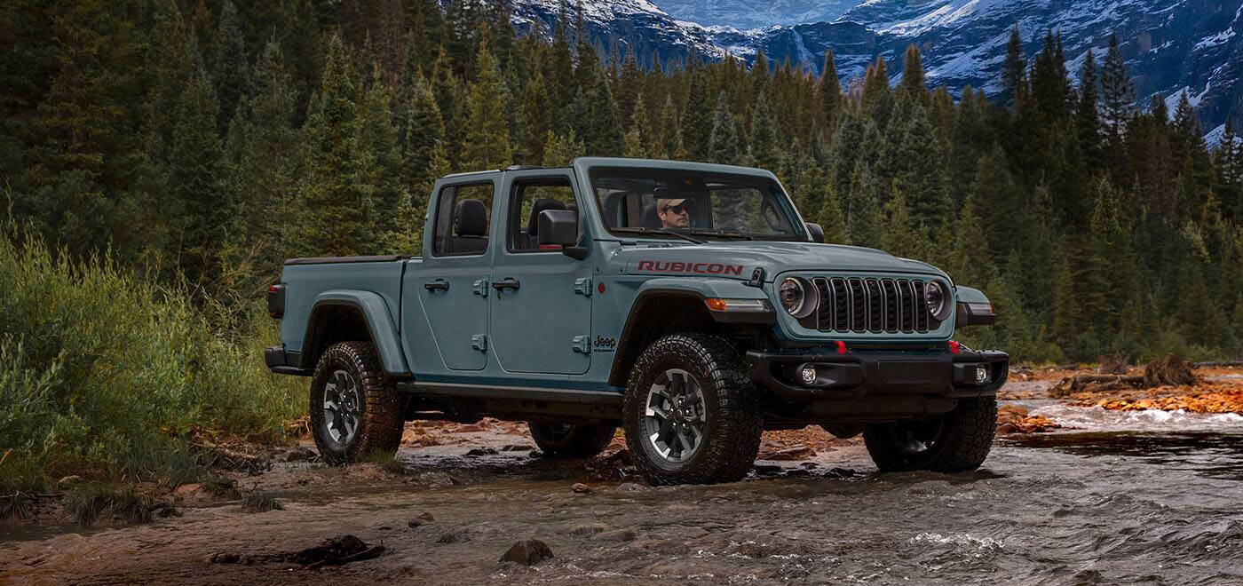 A passenger-side front angle of a gray 2026 Jeep Gladiator Rubicon X entering a stream in a mountain forest.