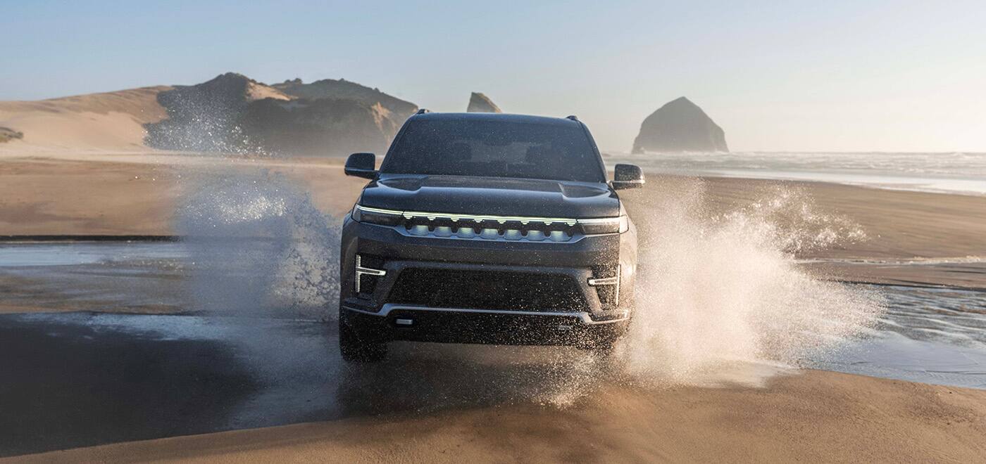 A head-on angle of a black 2026 Jeep Grand Wagoneer Upland traveling on a sandy beach at the water's edge at sunset, spraying water as it goes.