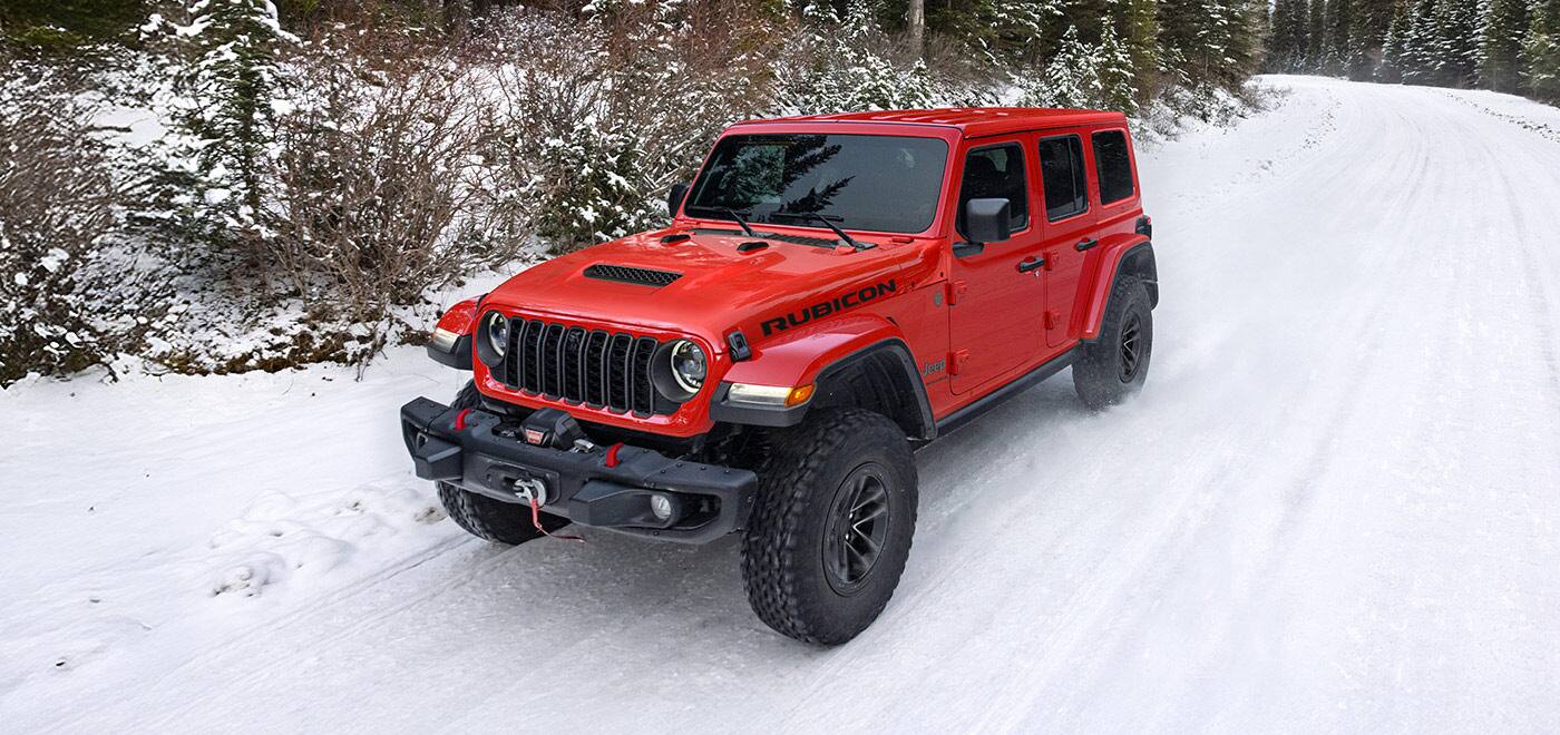 A red 2026 Jeep Wrangler Rubicon four-door traveling through a forest on a snow-covered road.