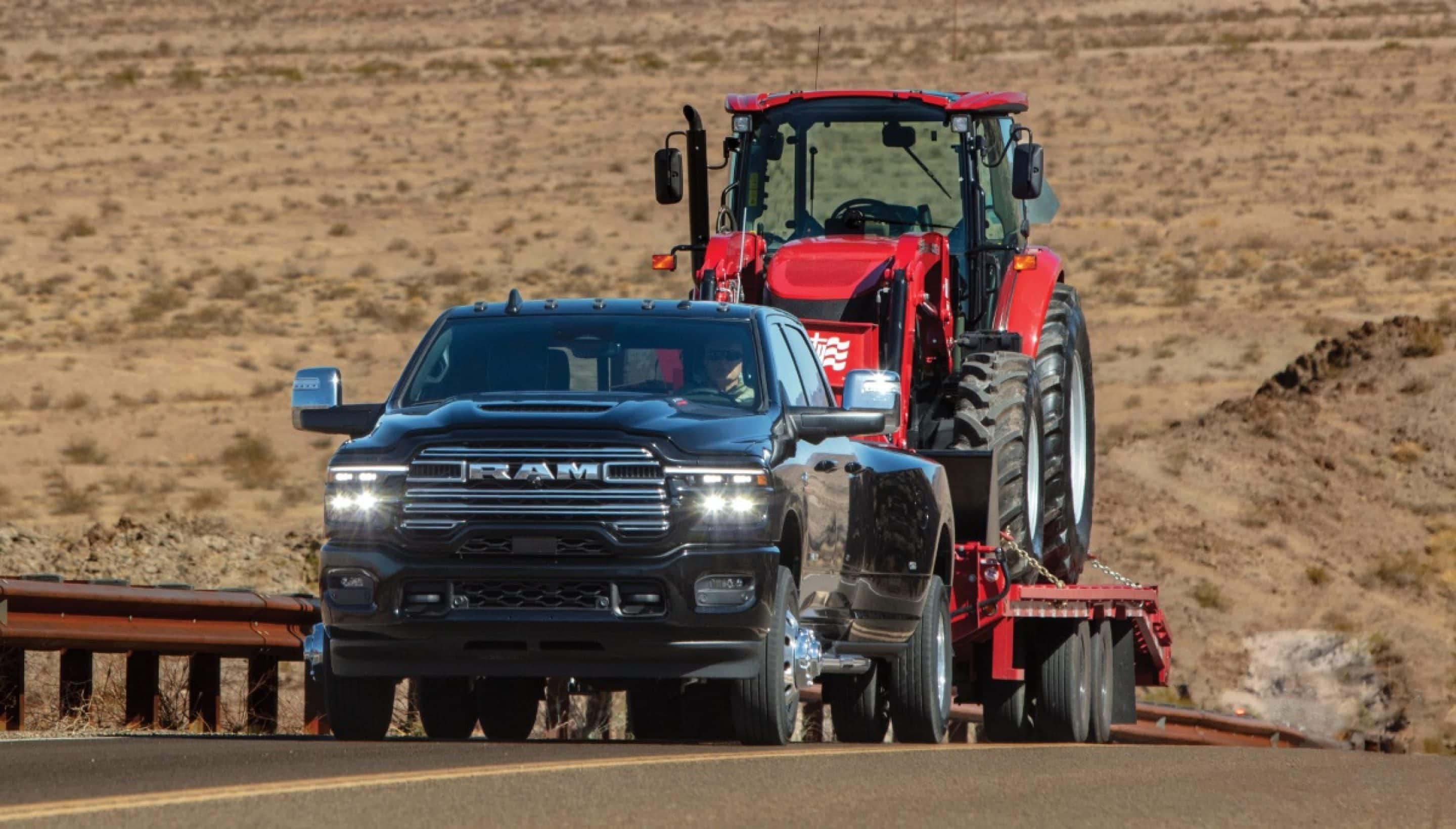 A black 2026 Ram 3500 Limited Longhorn 4x4 Crew Cab heavy duty truck hauling a large agriculture tractor on a flatbed trailer traveling down a rural highway.