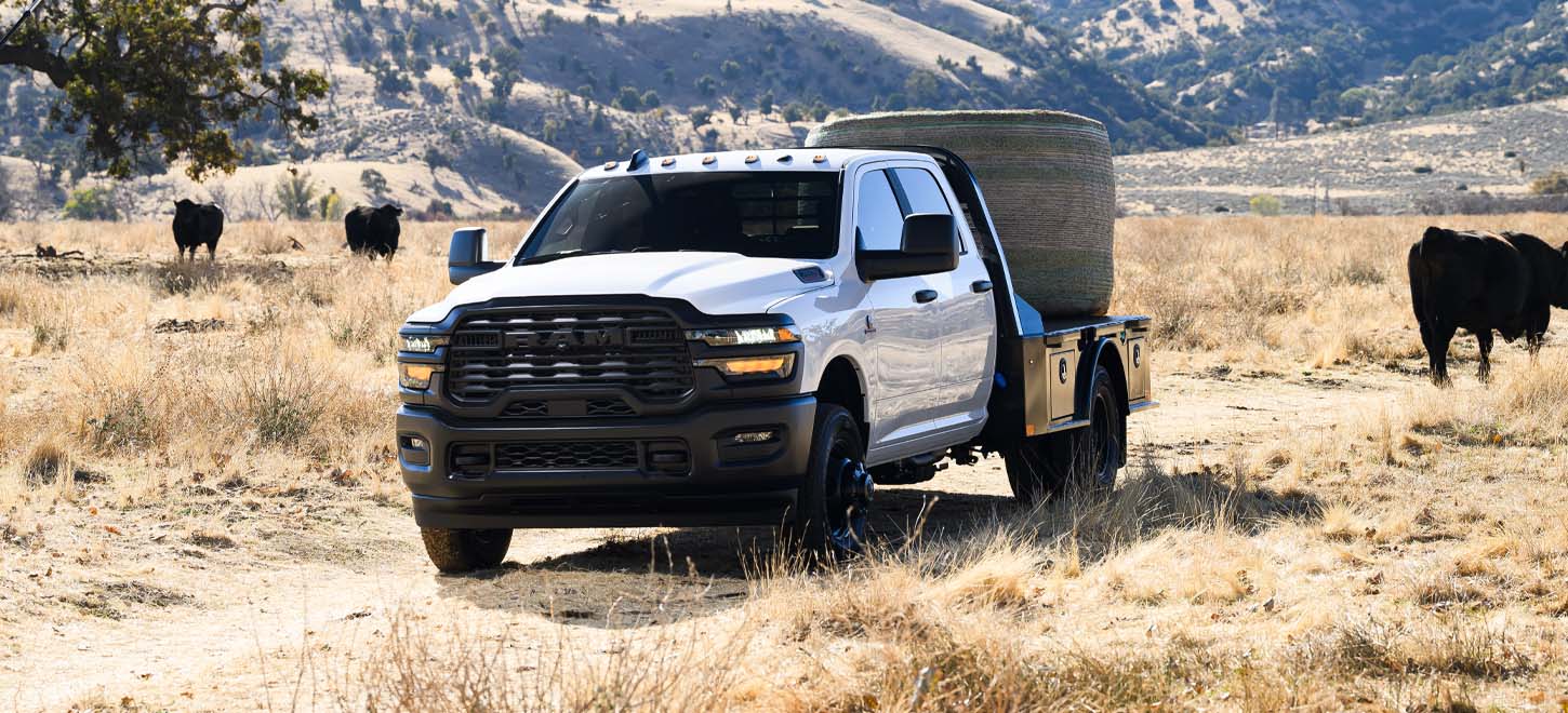 A white 2026 Ram 3500 Tradesman 4x2 Chassis Cab Crew Cab parked on a cattle ranch, carrying a bale of hay on a platform upfit.