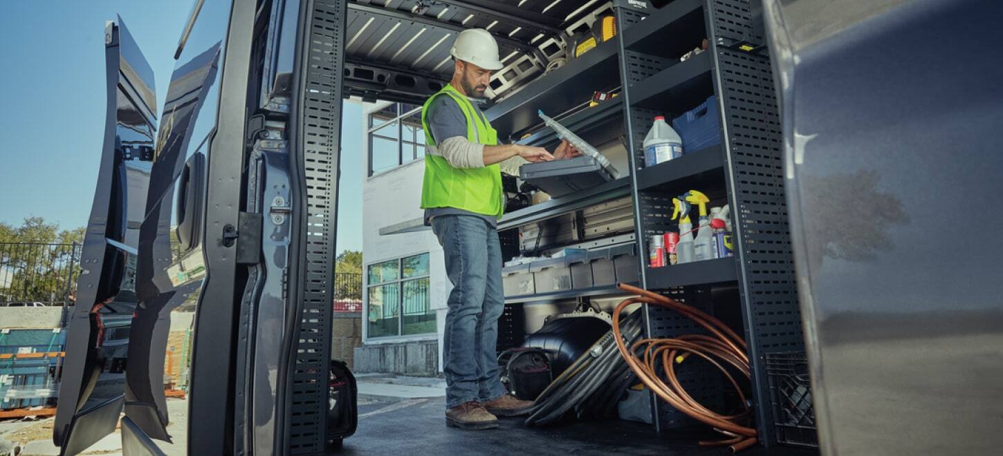 The 2026 Ram ProMaster with its passenger-side sliding door and rear doors open, displaying shelves full of tools and equipment and a technician working on a laptop.