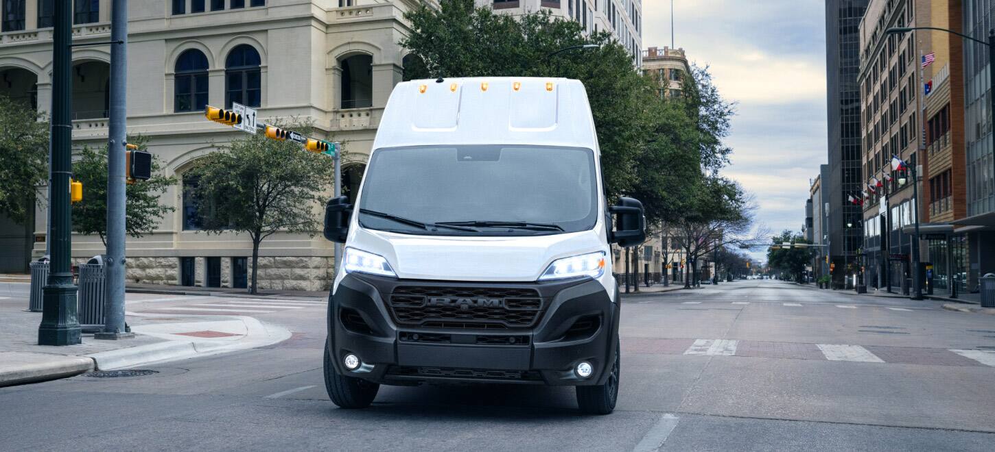 A head-on angle of a white 2026 Ram ProMaster 3500 SLT Plus Cargo Van Super High Roof, with Black Appearance Package,  traveling down a city street.