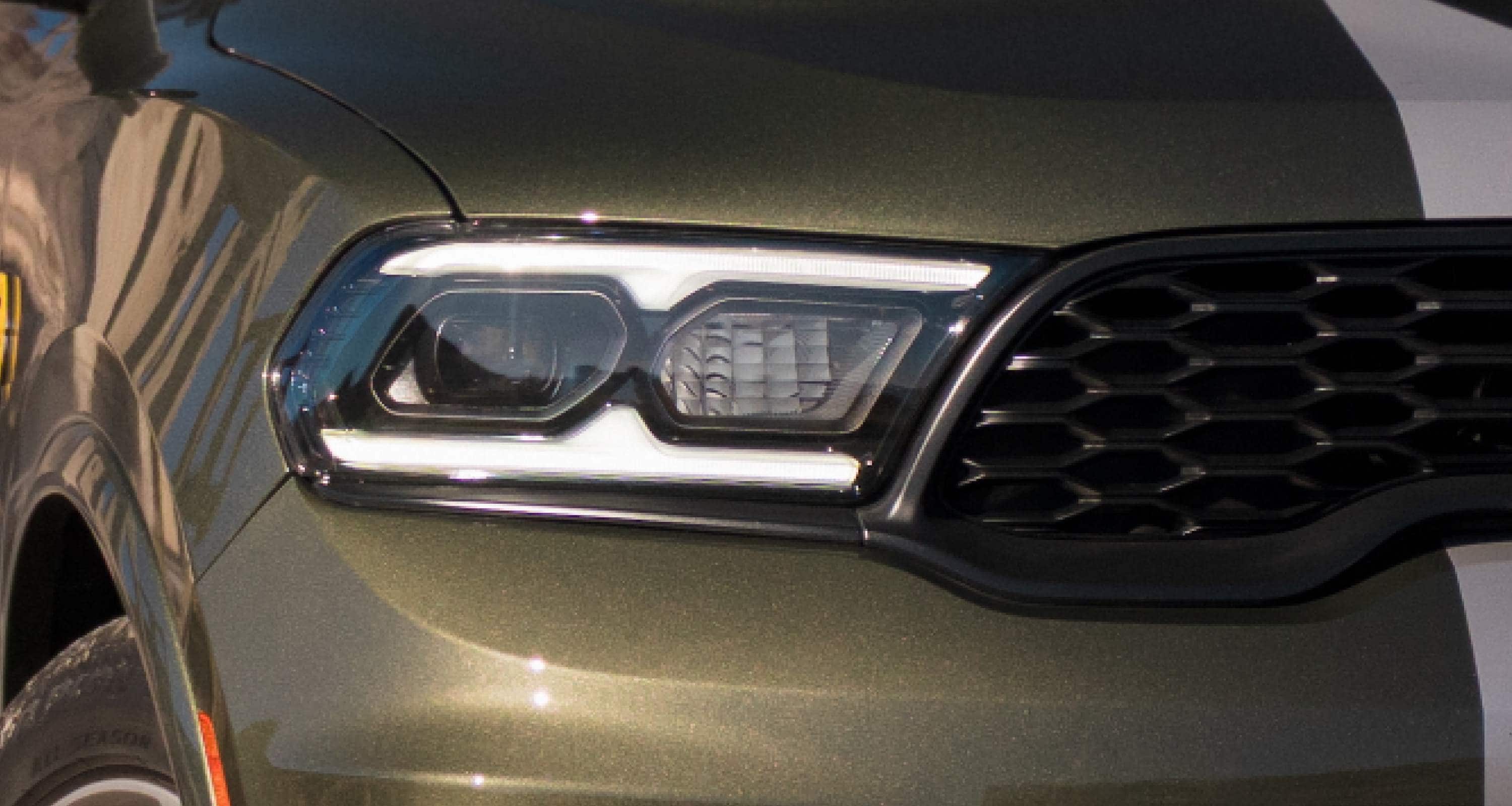 The grille and one of the headlamps on the passenger-side of an olive green 2027 Dodge Durango GT Hemi V8 with dual white center stripes.
