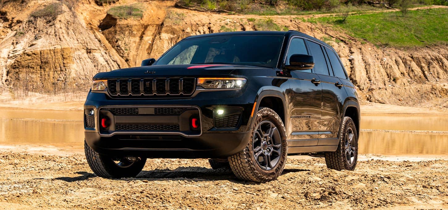 A black 2027 Jeep Grand Cherokee Trailhawk parked on a sandy trail beside a stream.