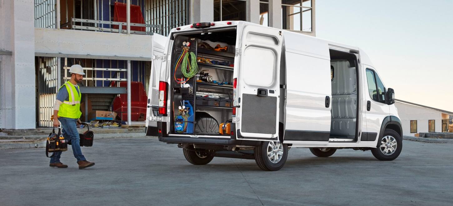 The rear of a white 2027 Ram ProMaster 2500 SLT High Roof Cargo Van with its passenger-side sliding door and rear doors open, displaying shelves full of tools and a technician walking toward the van.