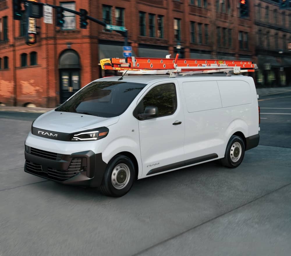 A driver-side front angle of a white 2027 Ram ProMaster City Tradesman Cargo Van traveling through an intersection on a city street.